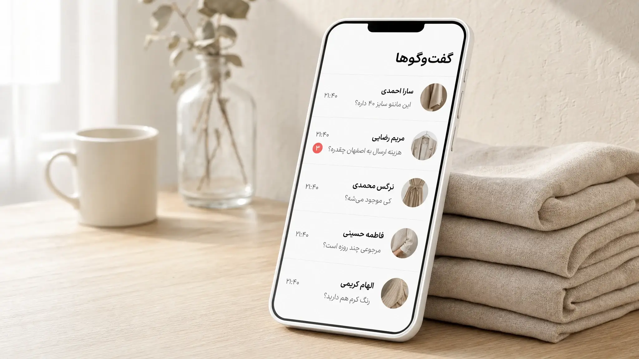 گوشی تکیه‌داده به پارچه کتان که فهرست گفت‌وگوهای واتساپ یک فروشگاه پوشاک را نشان می‌دهد؛ هر ردیف یک سوال خرید مثل سایز، هزینه ارسال و مرجوعی است و یکی از ردیف‌ها نشان قرمز پیام خوانده‌نشده دارد.